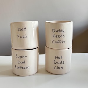 Puede incluir: Cuatro tazas de cerámica apiladas, de color blanquecino, con texto escrito a mano en negro. Las tazas dicen: "Dad Fuel", "Super Dad Espresso", "Daddy Needs Coffee" y "Hot Dads Club". Las tazas son cilíndricas y tienen un acabado liso y brillante.