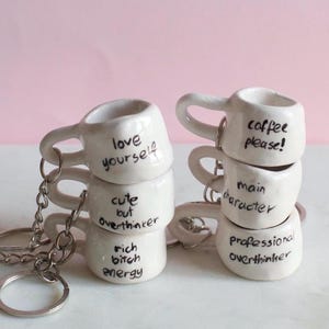 Pode incluir: Mini chaveiros de caneca de cerâmica branca com frases humorísticas em tinta preta. Uma pilha diz "love yourself", "cute but overthinker" e "rich bitch energy". A outra diz "coffee please!", "main character" e "professional overthinker".