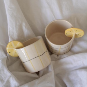 Puede incluir: Dos tazas de cerámica con un asa de rodaja de limón. Las tazas son de color blanco roto con rayas verticales amarillas y una línea horizontal negra. El asa de rodaja de limón es amarilla con puntos negros. Las tazas están sobre una tela blanca.
