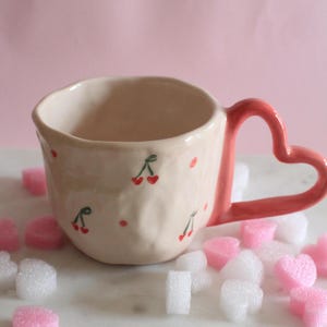 Puede incluir: Una taza de cerámica color crema con un asa en forma de corazón. La taza está decorada con pequeñas cerezas rojas y puntos. El asa y los puntos están pintados en un suave color rosa. La taza está rodeada de caramelos rosas y blancos en forma de corazón.