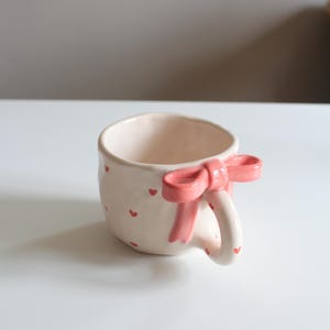 Puede incluir: Una taza de cerámica color crema con pequeños corazones rojos esparcidos por la superficie. El asa está adornada con un lazo rosa. Esta taza hecha a mano es perfecta para disfrutar de tu bebida caliente favorita.