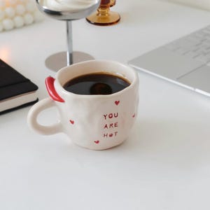 Può includere: Una tazza in ceramica color crema con una texture irregolare modellata a mano. La tazza presenta piccoli cuori rossi e la scritta rossa "YOU ARE HOT" stampata sul davanti. Un peperoncino rosso brillante è fissato sul lato vicino al manico.