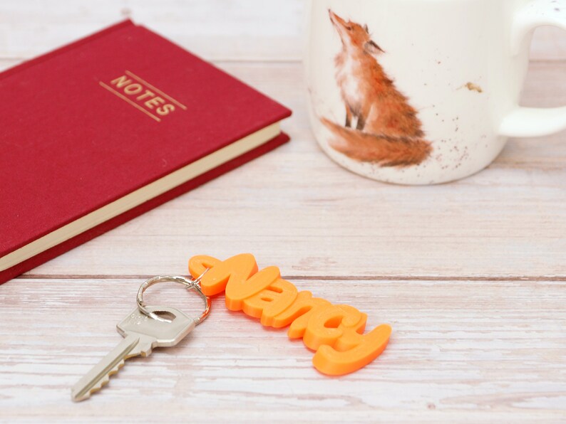 May include: An orange keychain spelling "Nancy" with a silver key attached, resting on a light wood surface. A red notebook with "NOTES" in gold lettering and a mug with a fox illustration are also present.