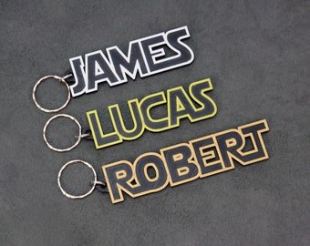 May include: Three keychains with black outlined lettering on a gray background. The top keychain is white with the name "JAMES", the middle keychain is yellow with the name "LUCAS", and the bottom keychain is gold with the name "ROBERT".