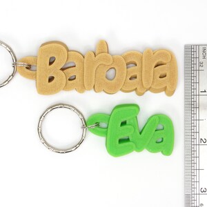 May include: Two keychains with names. One keychain says "Barbara" in tan, the other says "Eva" in green. Both have silver key rings. A ruler shows the keychains are approximately 3 inches long.