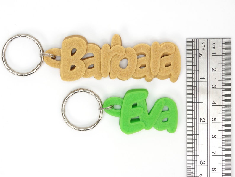 May include: Two keychains with names. One keychain says "Barbara" in tan, the other says "Eva" in green. Both have silver key rings. A ruler shows the keychains are approximately 3 inches long.