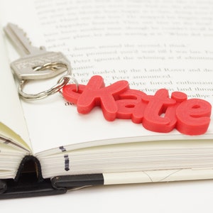 May include: A silver key with a key ring attached to a red keychain that spells out the name "Katie". The keychain rests on the open pages of a book.