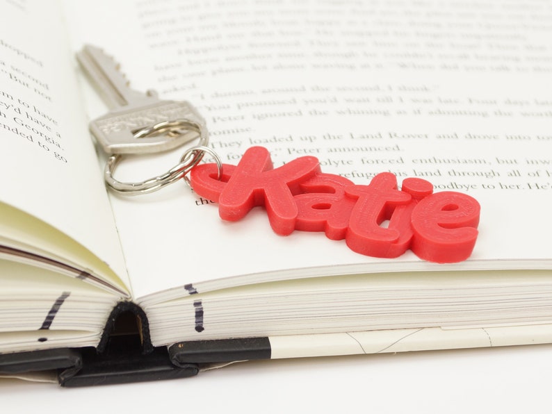 May include: A silver key with a key ring attached to a red keychain that spells out the name "Katie". The keychain rests on the open pages of a book.