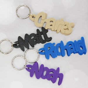 May include: Four personalized keychains in various colors: beige, black, blue, and purple. Each keychain features a name: 'Charles', 'Matt', 'Richard', and 'Noah' in a playful font, attached to a silver keyring. Ideal for gifts or personal use.