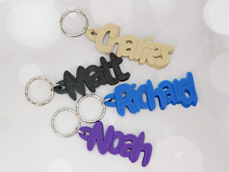 May include: Four personalized keychains in various colors: beige, black, blue, and purple. Each keychain features a name: 'Charles', 'Matt', 'Richard', and 'Noah' in a playful font, attached to a silver keyring. Ideal for gifts or personal use.