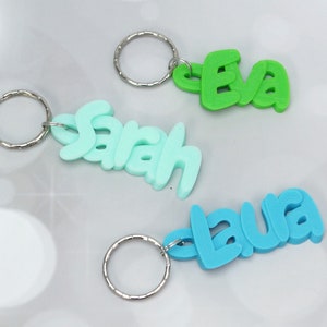 May include: Three colorful keychains with names: "Eva" in green, "Sarah" in light blue, and "Laura" in blue. Each keychain features a silver metal ring. The names are in a playful, bubble-style font.