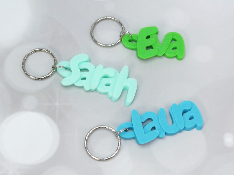 May include: Three colorful keychains with names: "Eva" in green, "Sarah" in light blue, and "Laura" in blue. Each keychain features a silver metal ring. The names are in a playful, bubble-style font.