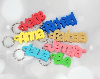 May include: Colorful name keychains in red, blue, yellow, tan, pink, and green. Each keychain features a different name in a playful, cursive font. The keychains are attached to silver keyrings, perfect for personalizing keys or bags.