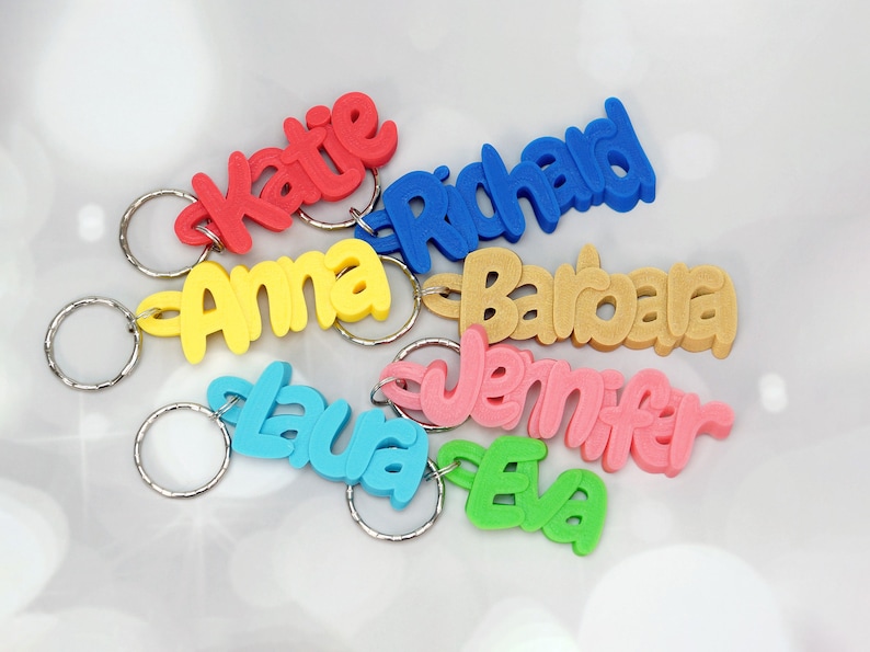 May include: Colorful name keychains in red, blue, yellow, tan, pink, and green. Each keychain features a different name in a playful, cursive font. The keychains are attached to silver keyrings, perfect for personalizing keys or bags.