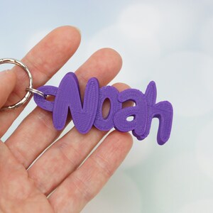 May include: A purple keychain with the name "Noah" in a stylized font. The keychain is attached to a silver metal ring. The background is a soft, blurred light blue and white.