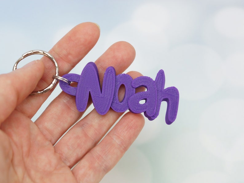 May include: A purple keychain with the name "Noah" in a stylized font. The keychain is attached to a silver metal ring. The background is a soft, blurred light blue and white.