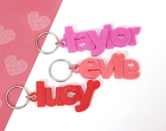 May include: Three personalized name keychains in shades of pink and red. The names "Taylor", "Evie", and "Lucy" are displayed in a playful font, with heart-shaped cutouts. Each keychain has a silver metal ring.