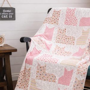 Könnte beinhalten: Ein Steppdecke mit einem sich wiederholenden Muster aus Katzensilhouetten in verschiedenen Rosatönen, Blumenmustern und gestreiften Stoffen. Die Decke liegt über einem schwarzen Holzschaukelstuhl, auf dem eine schwarz-weiße Katze sitzt.