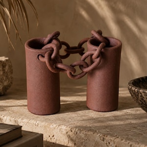 May include: A pair of cylindrical terracotta-colored vases connected by a decorative chain made of the same material. The vases have a matte, textured finish and are linked by several interlocking oval-shaped links that span the gap between them.
