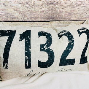 May include: Rectangular beige pillow with the numbers 71322 printed in distressed black font. The word "home" is written in cursive below the numbers. The pillow is on a white surface.