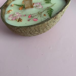 K&ouml;nnte beinhalten: Eine handgemachte Kerze in einer Kokosnussschale. Die Kerze hat hellgr&uuml;nes Wachs, verziert mit rosa Bl&uuml;ten, gr&uuml;nen Farnbl&auml;ttern und Goldflocken. Ein Holzdocht befindet sich in der Mitte. Die Schale ist braun und oval geformt.