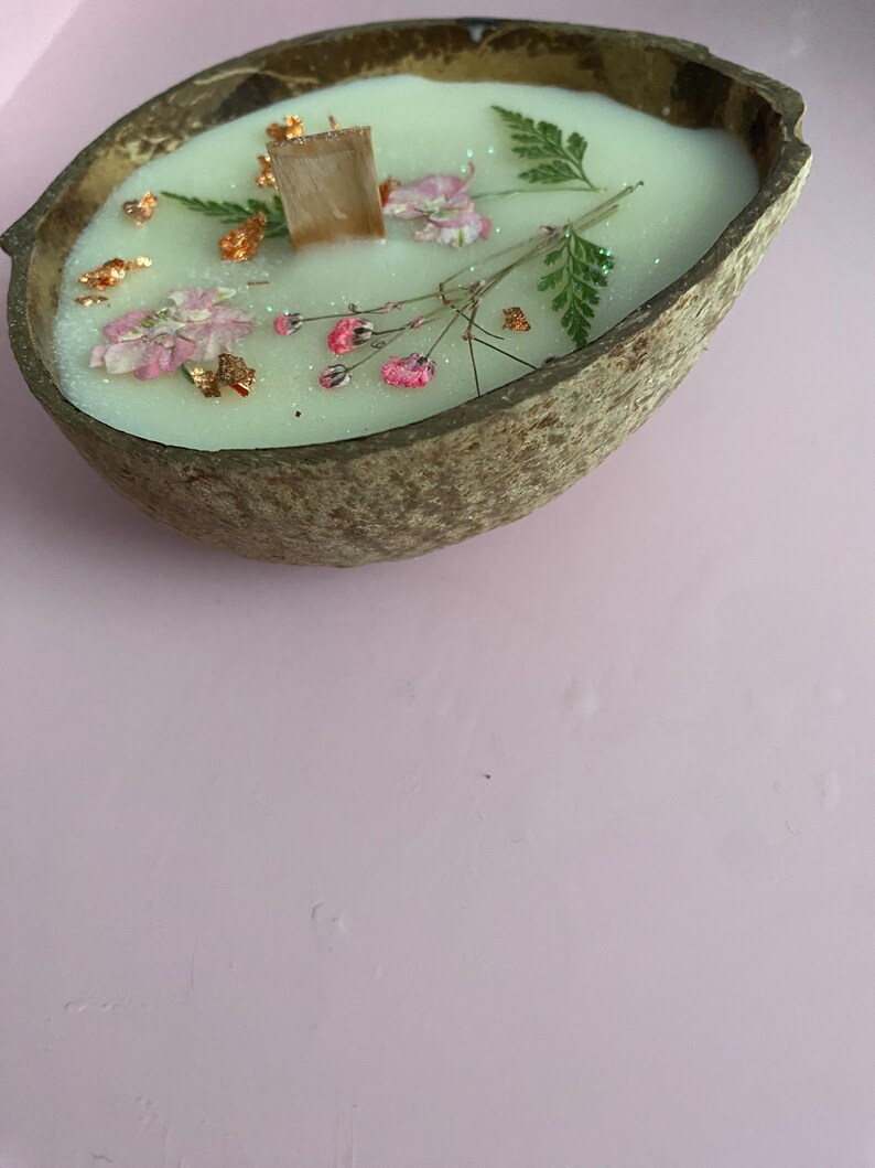 K&ouml;nnte beinhalten: Eine handgemachte Kerze in einer Kokosnussschale. Die Kerze hat hellgr&uuml;nes Wachs, verziert mit rosa Bl&uuml;ten, gr&uuml;nen Farnbl&auml;ttern und Goldflocken. Ein Holzdocht befindet sich in der Mitte. Die Schale ist braun und oval geformt.