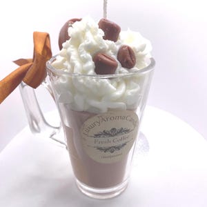 May include: A coffee-themed candle in a clear glass mug, topped with white whipped cream and chocolate-covered coffee beans. The candle has a brown liquid appearance and a label that reads "Luxury Aroma Candle Fresh Coffee Handpoured."
