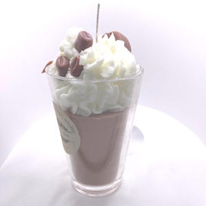 May include: A coffee-themed candle in a clear glass. The candle has a light brown wax base, topped with white whipped wax and brown coffee bean accents. A wick extends from the center. A small label is visible on the glass.