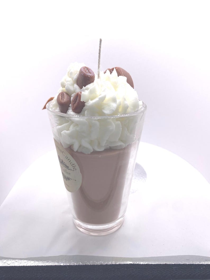 May include: A coffee-themed candle in a clear glass. The candle has a light brown wax base, topped with white whipped wax and brown coffee bean accents. A wick extends from the center. A small label is visible on the glass.