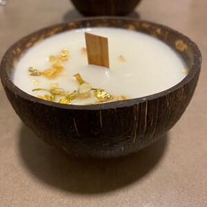 K&ouml;nnte beinhalten: Eine wei&szlig;e Kerze in einer dunkelbraunen Kokosnussschale mit einem Holzdocht. Die Kerze hat Goldflocken und kleine getrocknete Blumen auf der Oberseite. Eine weitere Kerze in einer Kokosnussschale ist im Hintergrund zu sehen.