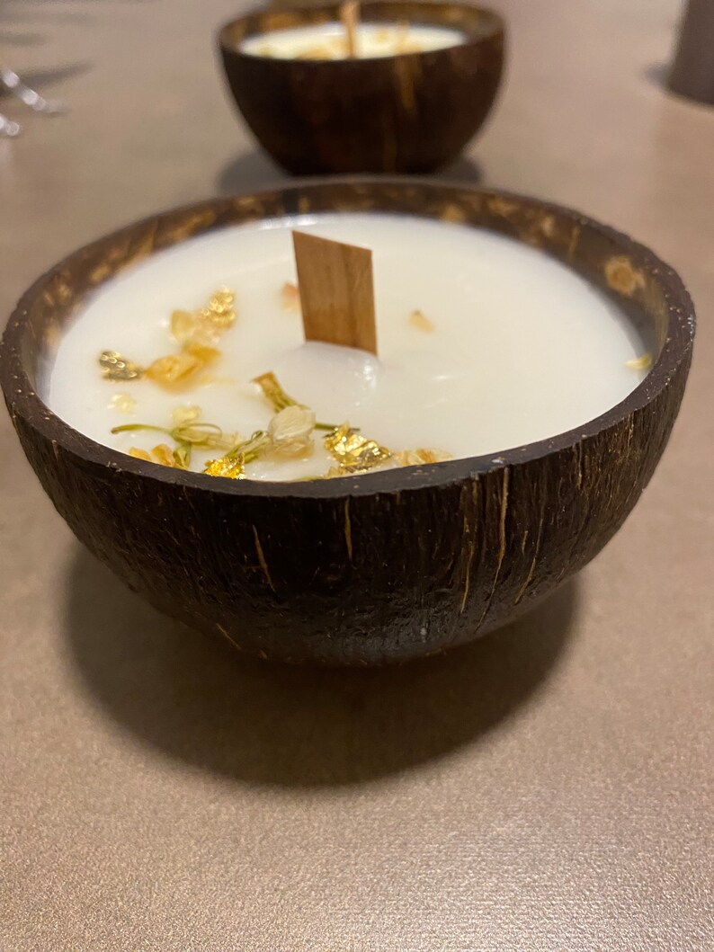 K&ouml;nnte beinhalten: Eine wei&szlig;e Kerze in einer dunkelbraunen Kokosnussschale mit einem Holzdocht. Die Kerze hat Goldflocken und kleine getrocknete Blumen auf der Oberseite. Eine weitere Kerze in einer Kokosnussschale ist im Hintergrund zu sehen.