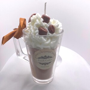 May include: A coffee-scented candle in a clear glass mug. The candle is brown, topped with white wax, and decorated with brown wax coffee beans. A brown ribbon is tied to the mug's handle. The label reads "Luxury Aroma Candle Fresh Coffee Handpoured."