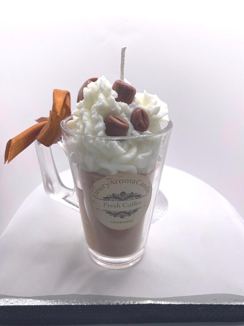 May include: A coffee-scented candle in a clear glass mug. The candle is brown, topped with white wax, and decorated with brown wax coffee beans. A brown ribbon is tied to the mug's handle. The label reads "Luxury Aroma Candle Fresh Coffee Handpoured."