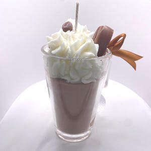 May include: A candle shaped like a coffee drink in a clear glass. The candle has a brown base, topped with white whipped cream, a chocolate accent, and a brown ribbon. A small brown bead and a wick are on top.