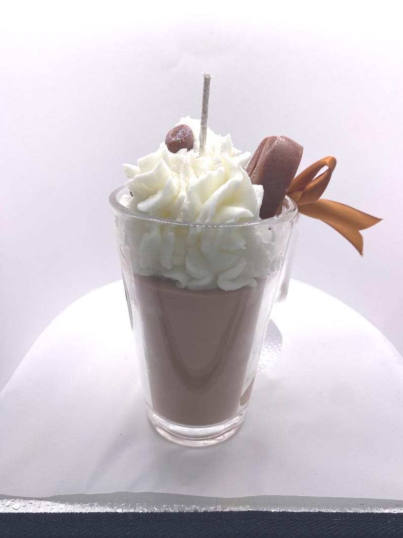 May include: A candle shaped like a coffee drink in a clear glass. The candle has a brown base, topped with white whipped cream, a chocolate accent, and a brown ribbon. A small brown bead and a wick are on top.