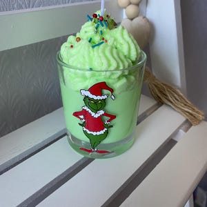 May include: A green candle in a clear glass, decorated with a Grinch sticker. The candle has a whipped wax top with colourful sprinkles and a white candle wick. The candle is on a white wooden surface.