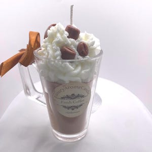 May include: A coffee-themed candle in a clear glass mug. The candle is brown, topped with white wax, and decorated with brown wax coffee beans. A brown ribbon is tied to the mug's handle. The label reads "Luxury Aroma Candle Fresh Coffee Handpoured."
