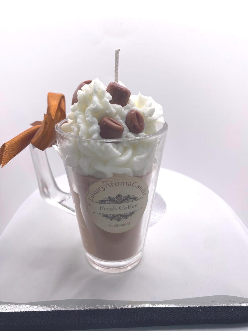 May include: A coffee-themed candle in a clear glass mug. The candle is brown, topped with white wax, and decorated with brown wax coffee beans. A brown ribbon is tied to the mug's handle. The label reads "Luxury Aroma Candle Fresh Coffee Handpoured."