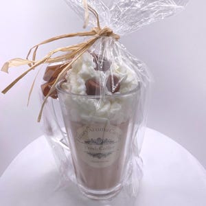 May include: A coffee-scented candle in a clear glass, resembling a coffee drink, topped with white wax and chocolate-colored wax pieces. The candle is wrapped in clear cellophane and tied with raffia. The label reads "Luxury Aroma Candle Fresh Coffee Handpoured."