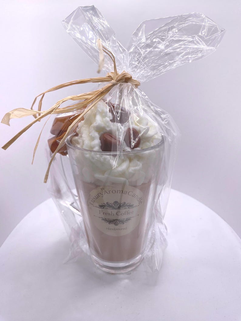 May include: A coffee-scented candle in a clear glass, resembling a coffee drink, topped with white wax and chocolate-colored wax pieces. The candle is wrapped in clear cellophane and tied with raffia. The label reads "Luxury Aroma Candle Fresh Coffee Handpoured."