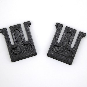 Könnte beinhalten: Zwei schwarze Plastikclips mit U-förmigem Design. Die Clips sind dazu bestimmt, Gegenstände zusammenzuhalten.