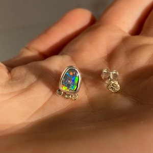 Puede incluir: Un par de pendientes de plata con una sola piedra de &oacute;palo en cada uno. El &oacute;palo es de color gris oscuro con destellos de verde, azul y rojo. Los pendientes est&aacute;n engastados con peque&ntilde;as cuentas doradas alrededor del &oacute;palo.