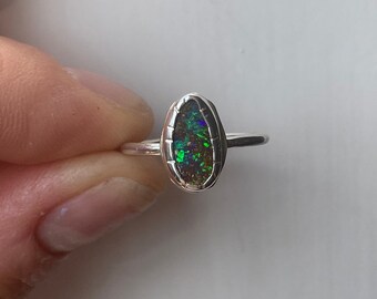 Puede incluir: Anillo de plata con una piedra de ópalo ovalada. El ópalo muestra colores verdes, azules y morados sobre un fondo oscuro. El anillo tiene una banda delgada de plata. El ópalo está engastado en plata.
