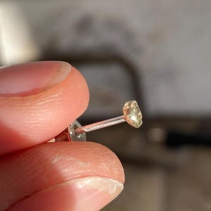 Puede incluir: Un pendiente dorado con una peque&ntilde;a piedra de forma irregular. El pendiente est&aacute; en un poste de plata.