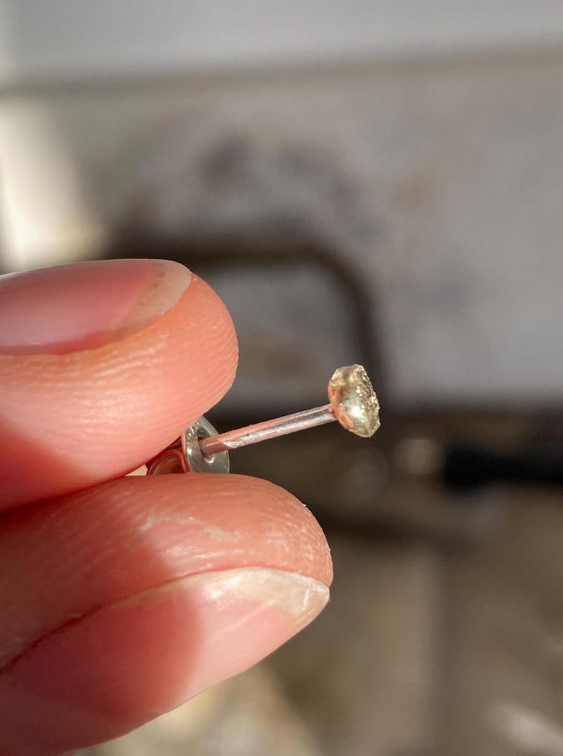 Puede incluir: Un pendiente dorado con una peque&ntilde;a piedra de forma irregular. El pendiente est&aacute; en un poste de plata.
