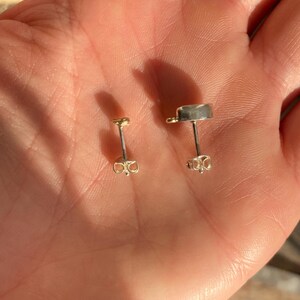 Puede incluir: Dos pares de pendientes, uno dorado y otro plateado, con un dise&ntilde;o sencillo de tachuela. Los pendientes se muestran en una mano.