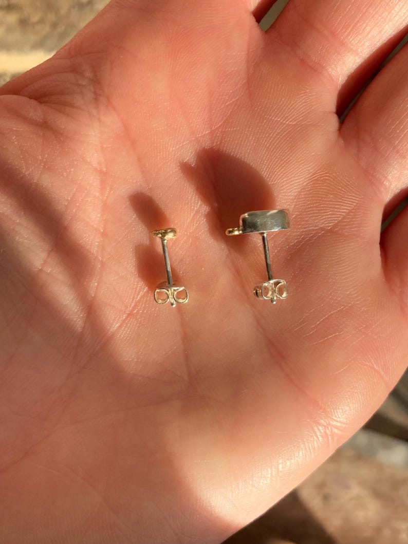 Puede incluir: Dos pares de pendientes, uno dorado y otro plateado, con un dise&ntilde;o sencillo de tachuela. Los pendientes se muestran en una mano.