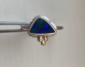 Puede incluir: Un anillo de plata con una piedra preciosa de ópalo negro triangular engastada en una banda de plata texturizada. El anillo tiene tres pequeñas bolas de oro en la parte inferior.