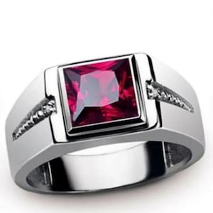 May include: A silver-toned ring featuring a large, square-cut, deep red gemstone set in the center. The band has a polished metallic finish and includes small, clear accent stones embedded along the sides of the central setting.