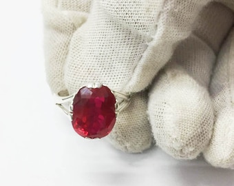 Peut inclure: Une bague en argent ornée d'une grosse pierre précieuse rouge foncé de taille ovale. La pierre précieuse est au centre, sertie dans un délicat anneau en argent avec un design à tige fendue. La bague est tenue par une main gantée sur fond blanc.